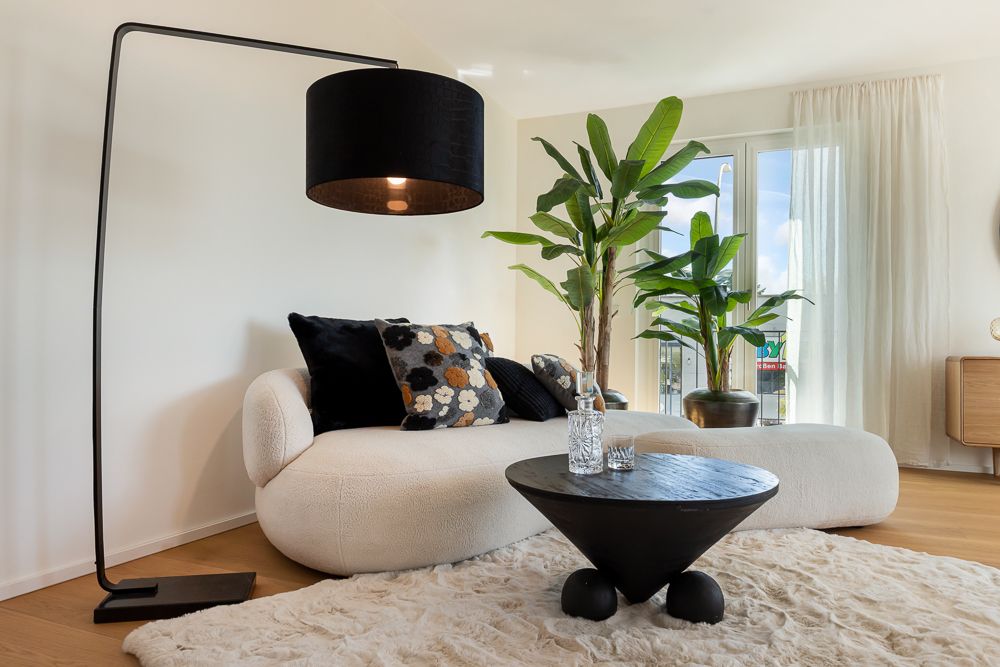 Modernes Wohnzimmer mit weißem Sofa, schwarzen Kissen, Stehlampe, Pflanzen und rundem Couchtisch.