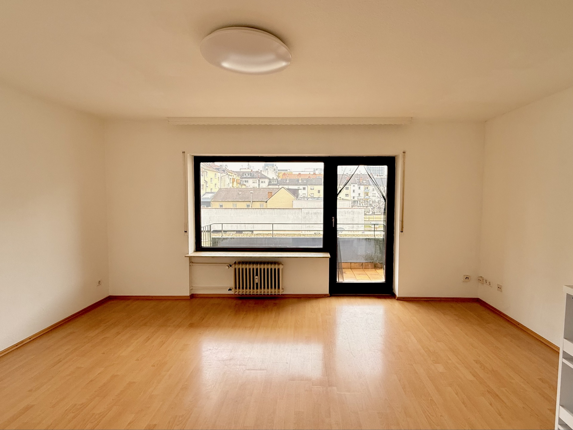 Leeres Zimmer mit Holzfußboden, großem Fenster und Tür zum Balkon.