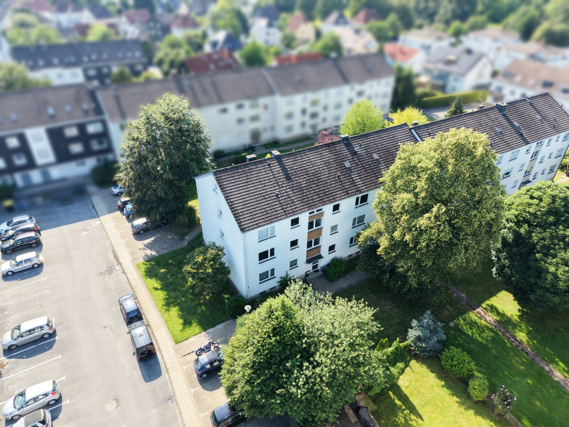 Mehrfamilienhaus mit grünem Vorgarten, Bäumen und angrenzendem Parkplatz aus der Vogelperspektive.