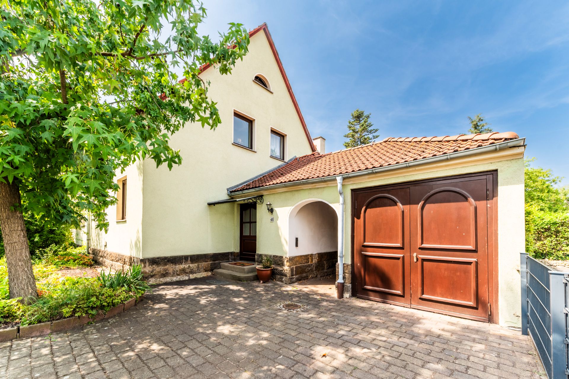 Einfamilienhaus mit gelber Fassade, rotem Dach und brauner Garage, davor gepflasterter Hof.
