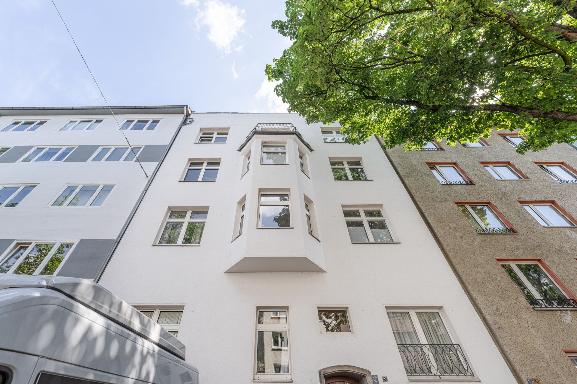 Weiße Wohnhausfassade mit Erker und mehreren Fenstern, daneben ein Baum.