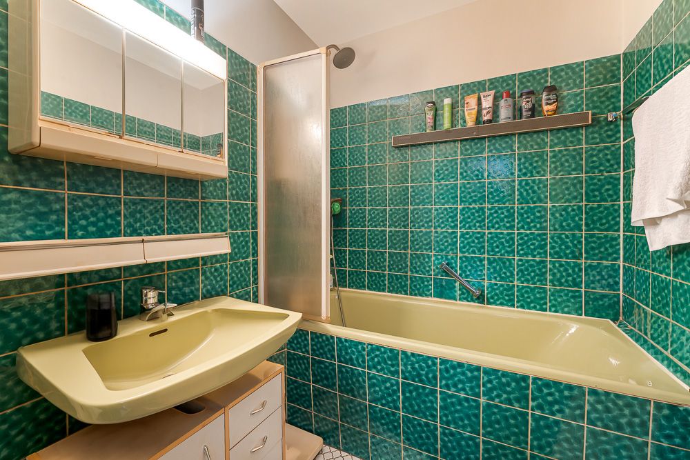 Badezimmer mit grünen Fliesen, gelber Badewanne, Waschbecken und Wandregal mit Pflegeprodukten.