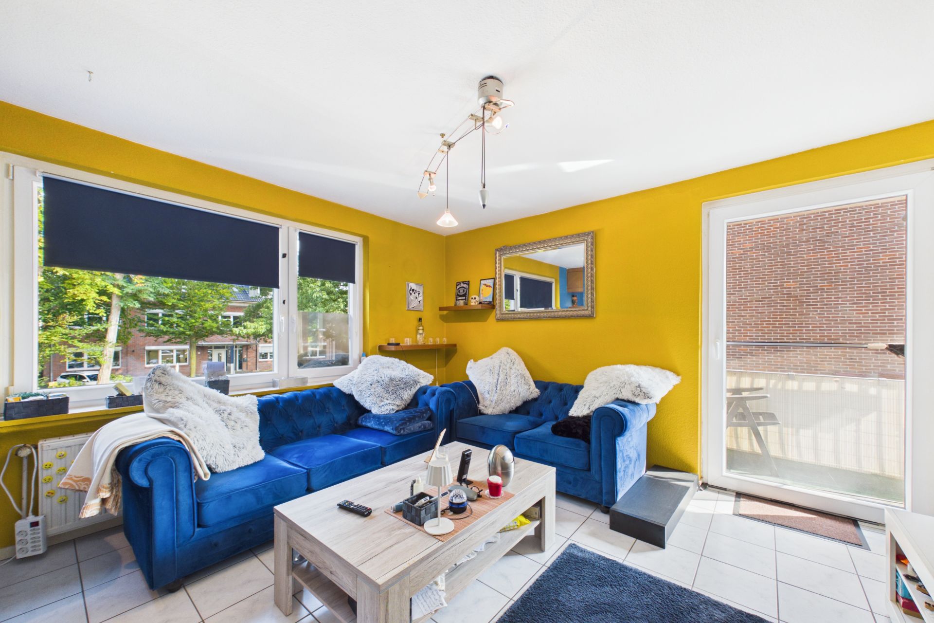 Wohnzimmer mit gelber Wand, blauen Sofas, Couchtisch und großem Fenster mit Blick nach draußen.