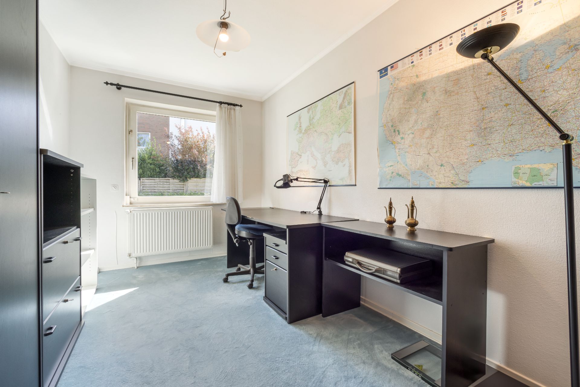 Helles Arbeitszimmer mit Schreibtisch, Stuhl, Regal und großen Landkarten an der Wand.
