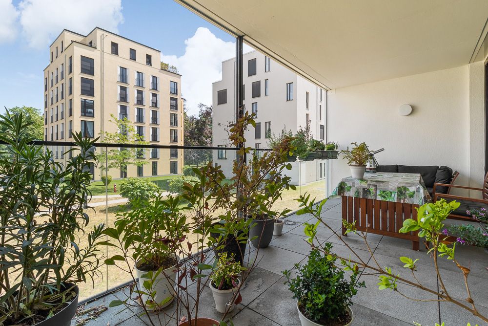 Großer Balkon mit vielen Pflanzen, Sitzgruppe und Blick auf moderne Wohngebäude.