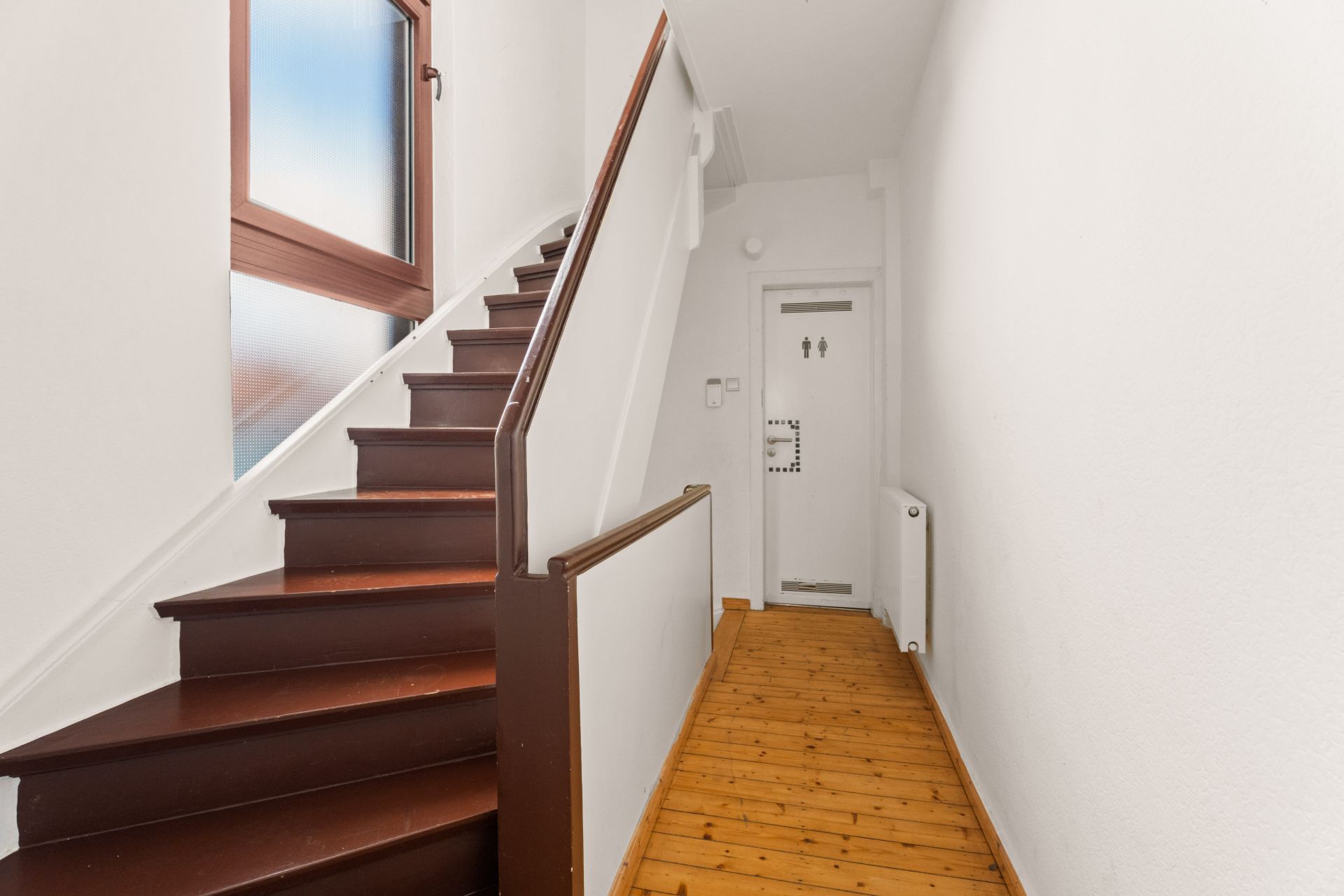 Flur mit brauner Holztreppe, Fenster und weißer Tür am Ende.