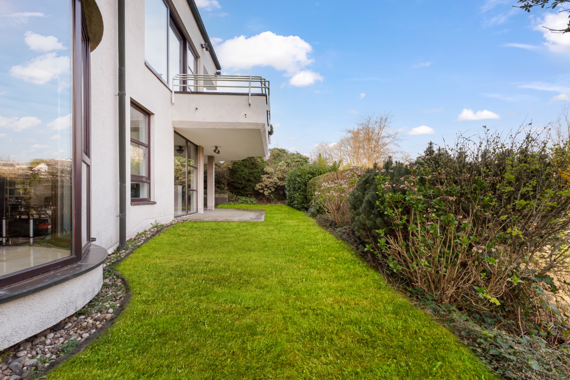 Grüner Garten mit Terrasse neben einem modernen Wohnhaus, umgeben von Büschen.