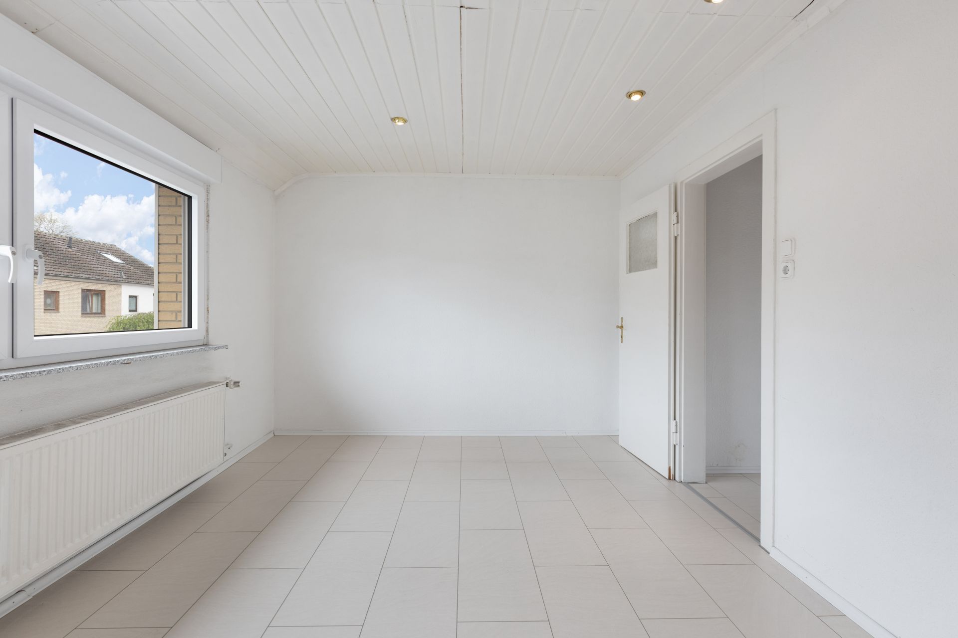 Heller, leerer Raum mit Fliesenboden, weißer Decke, Fenster und geöffneter Tür.