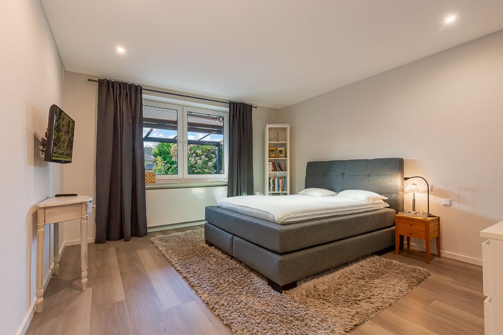 Modernes Schlafzimmer mit grauem Bett, Teppich, Nachttisch, Regal und großem Fenster.