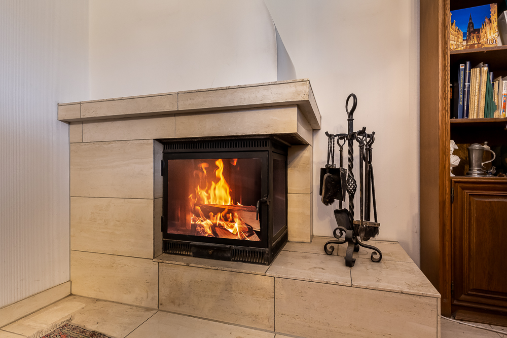 Ein moderner Kamin mit brennendem Feuer, daneben Kaminbesteck und ein Bücherregal.