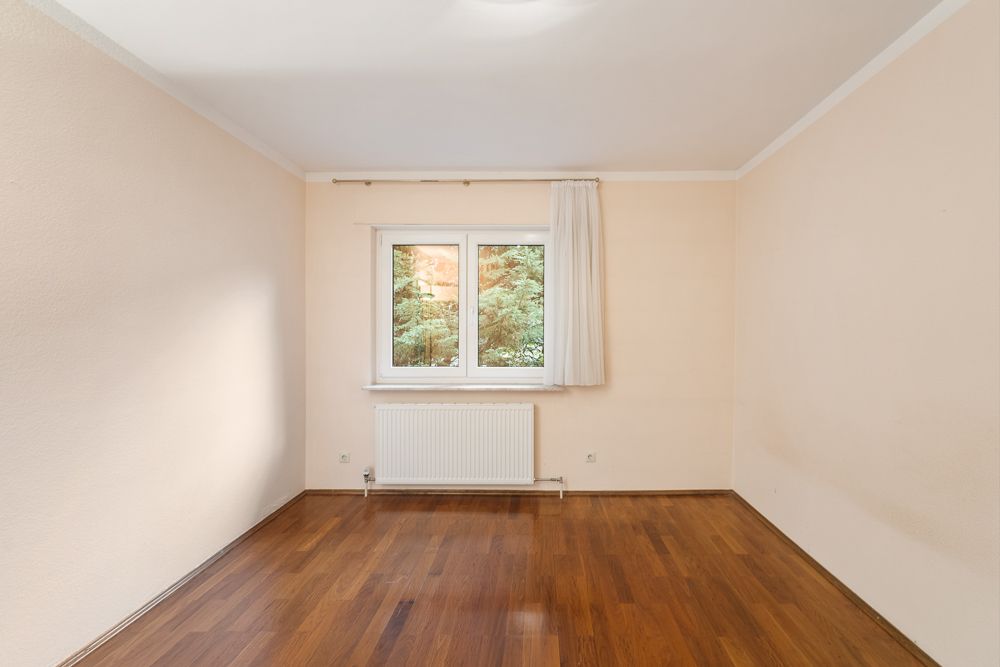 Leerstehender Raum mit Holzfußboden, cremefarbenen Wänden und einem Fenster mit Vorhang.