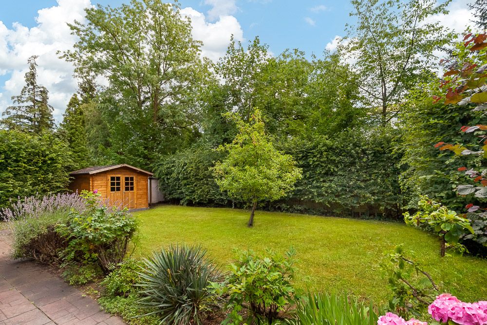 Garten mit Rasen, Baum, Blumenbeeten und einem kleinen Holzhaus im Hintergrund.