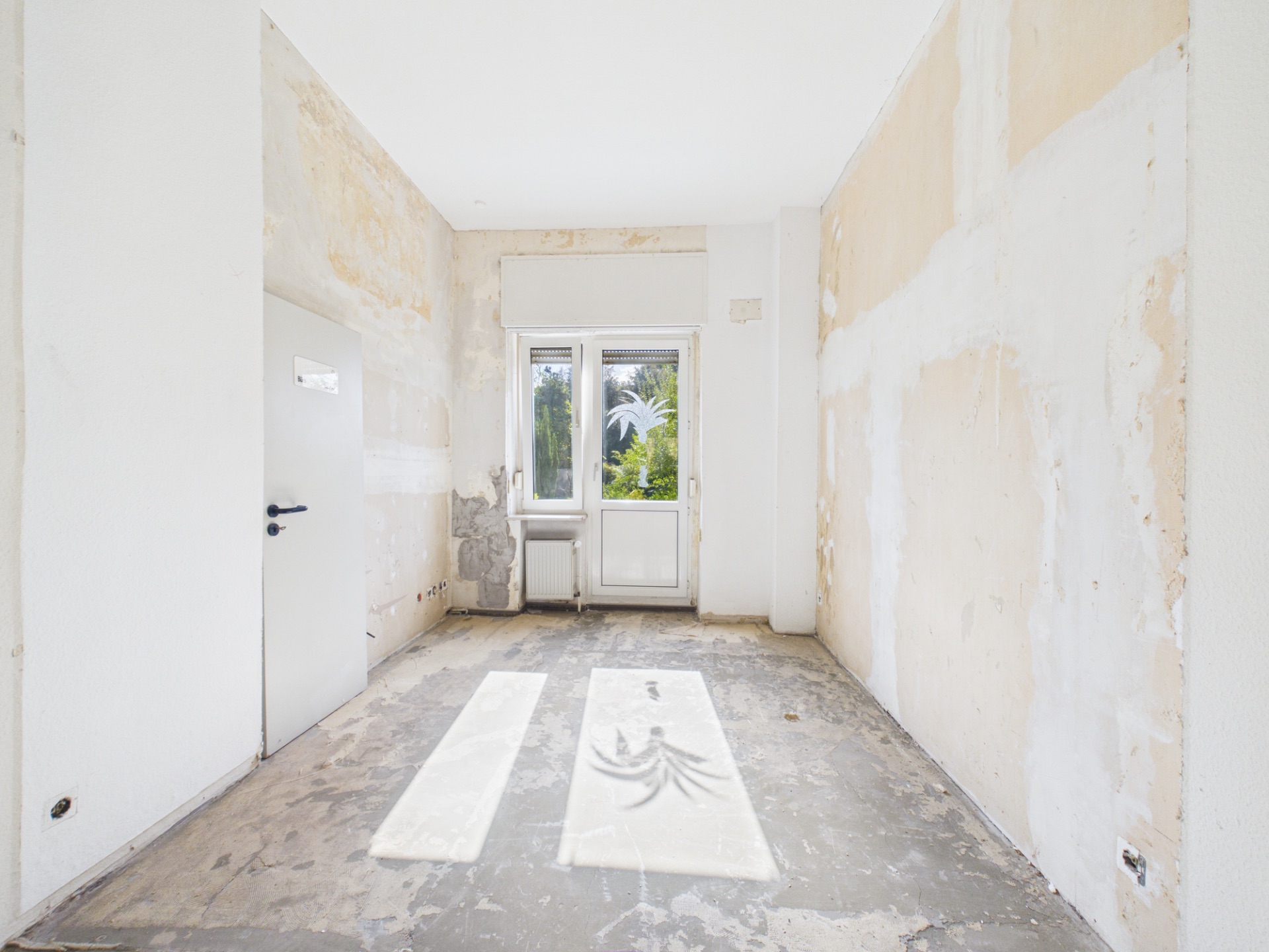 Leerer Raum mit ungepflegten Wänden, offenem Estrichboden und Fenster mit Blick ins Grüne.