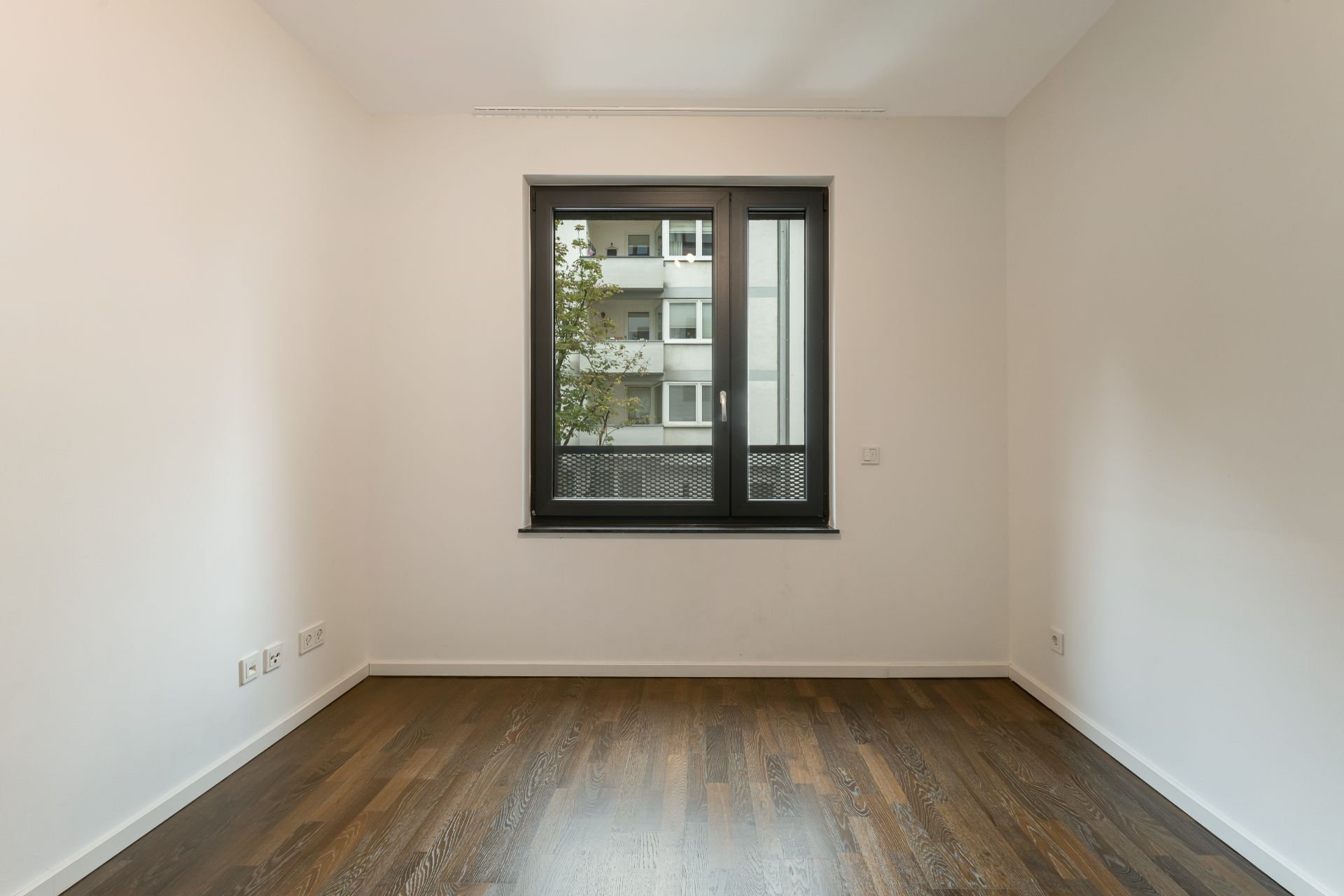 Leerer Raum mit dunklem Holzboden und Fenster, durch das ein Wohnhaus zu sehen ist.