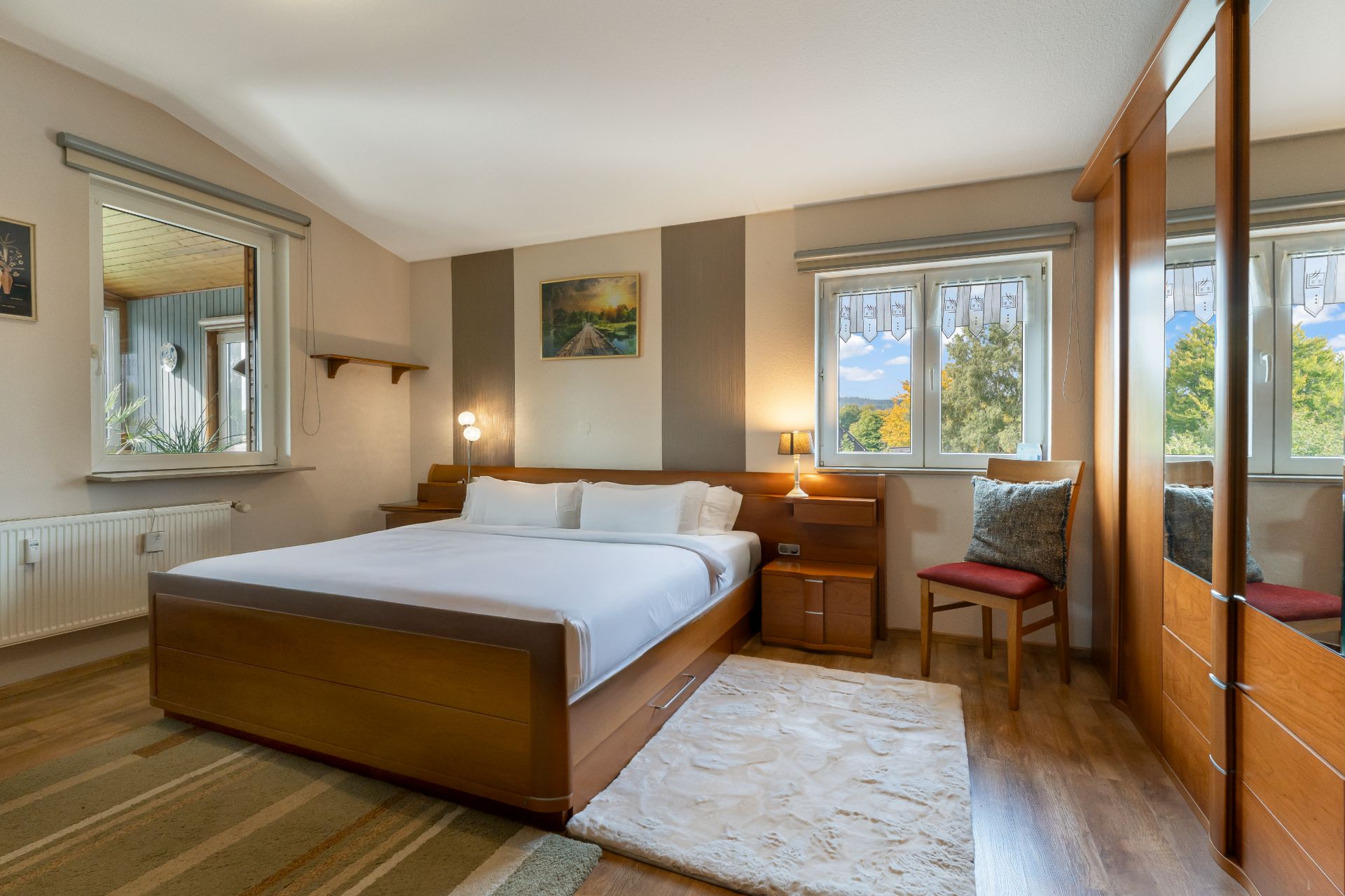 Helles Schlafzimmer mit großem Holzbett, weißen Kissen, Teppich, Stuhl und Spiegel-Kleiderschrank.