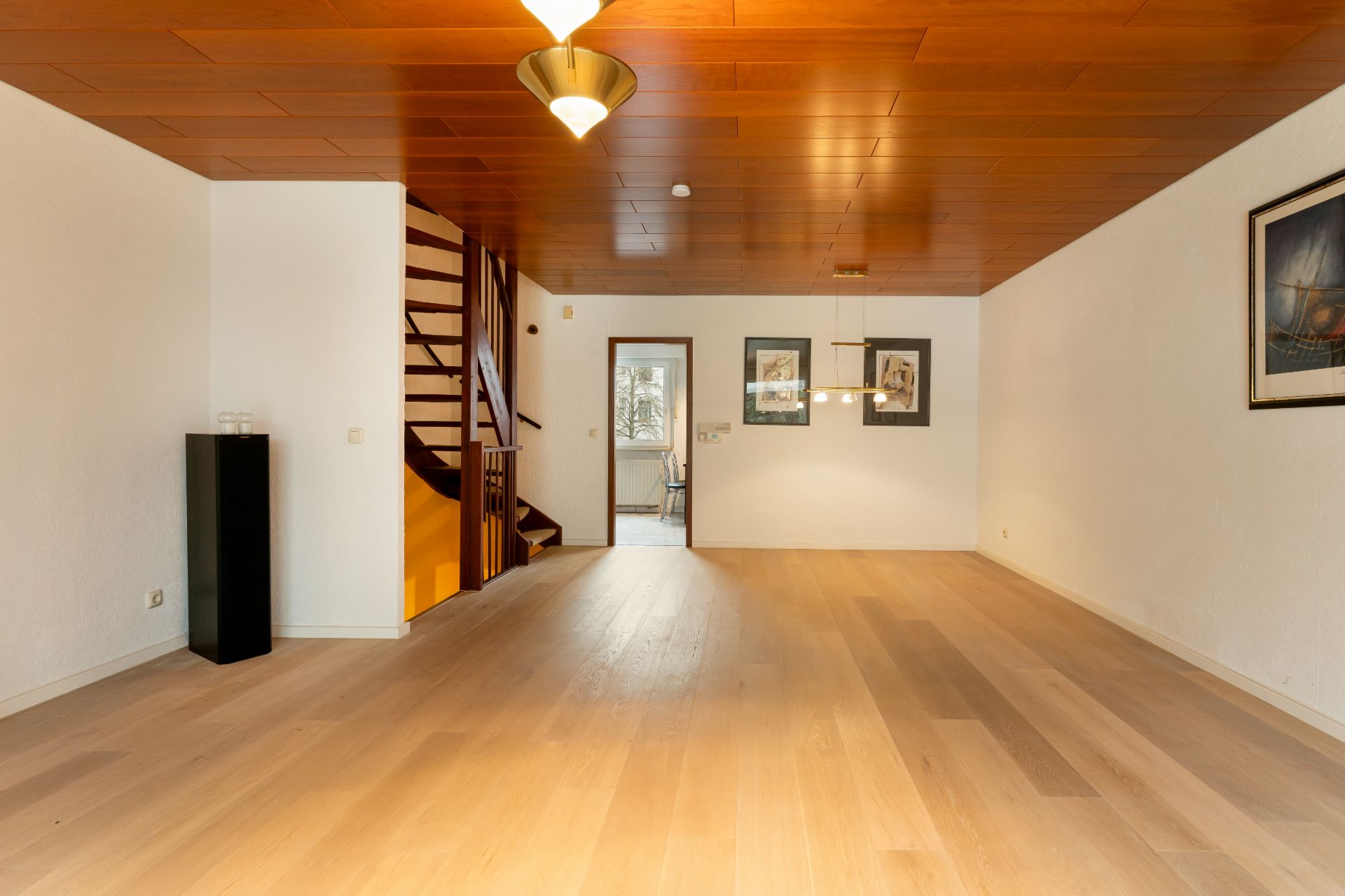 Leeres Wohnzimmer mit Holzdecke, Parkettboden, Treppe und Bildern an der Wand.