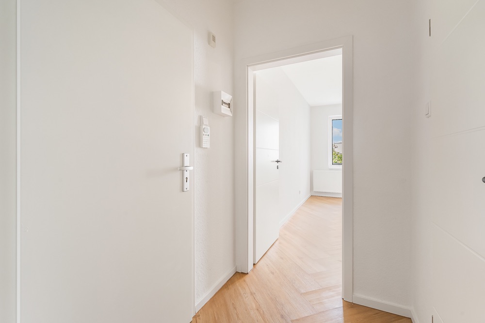 Heller Flur mit Holzboden und weißer Tür, Blick in ein Zimmer mit Fenster.