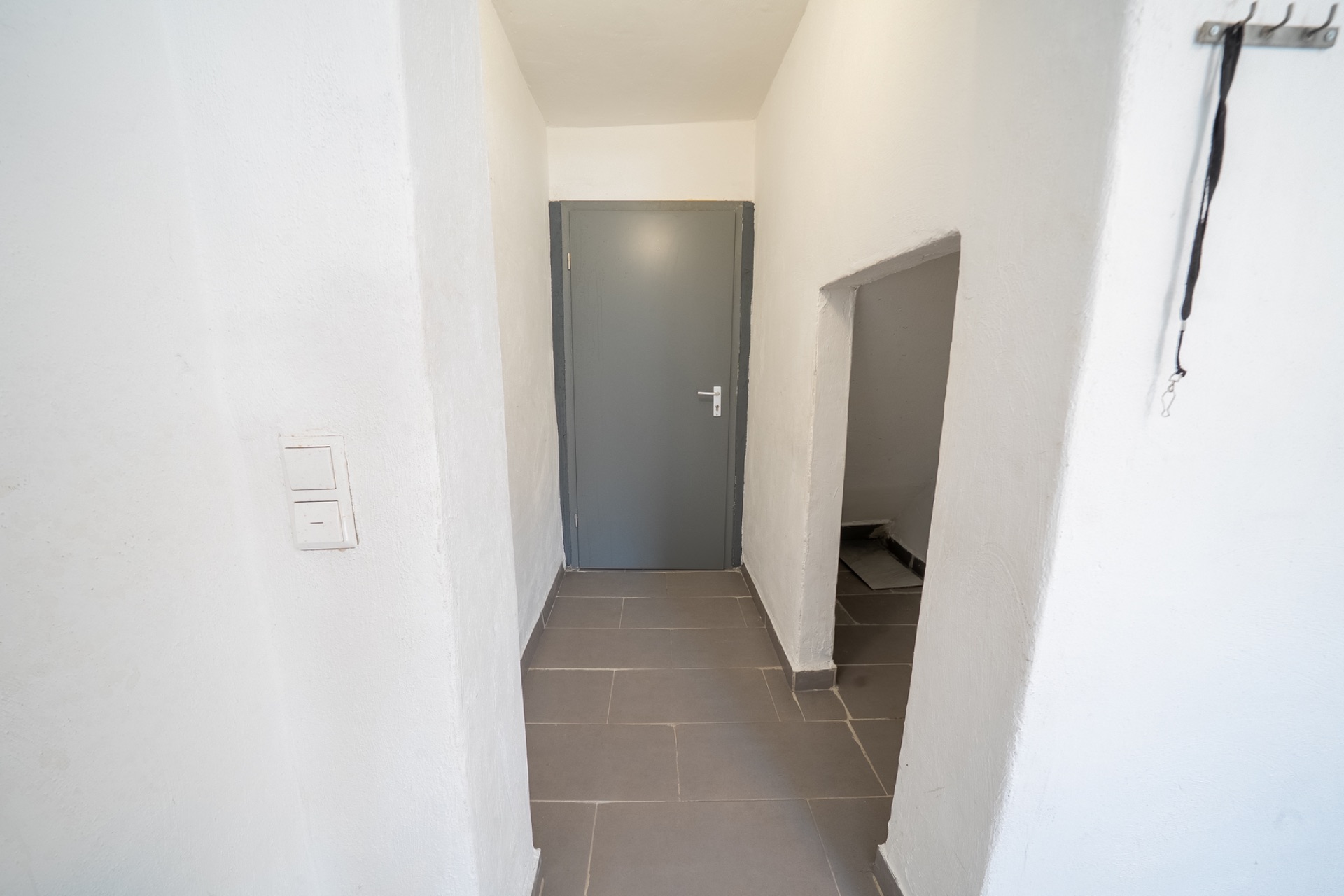 Heller Flur mit grauer Tür, weißen Wänden und einem kleinen Durchgang rechts.