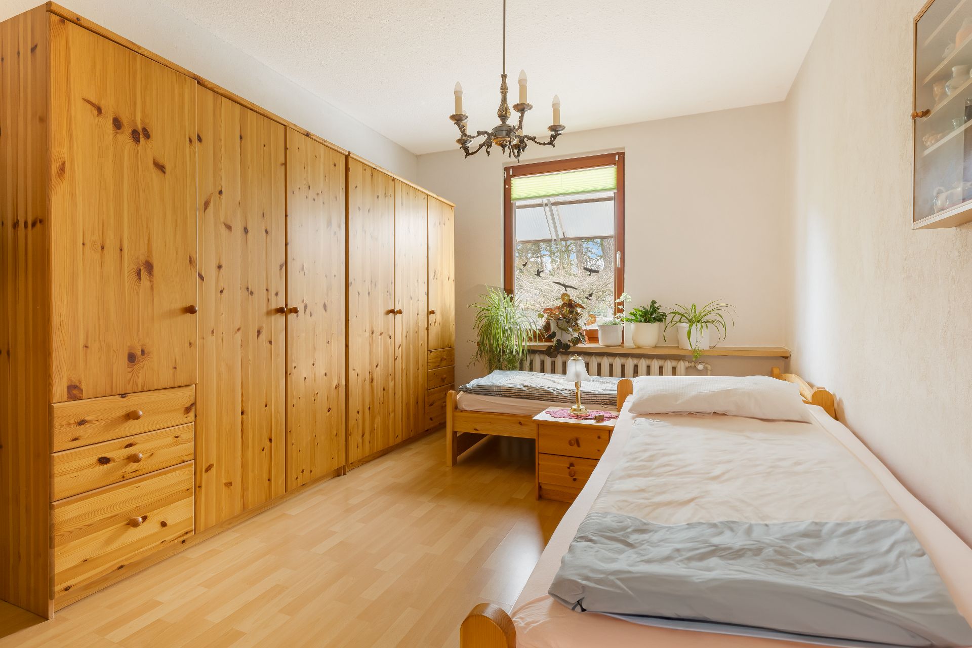 Helles Schlafzimmer mit zwei Einzelbetten, Holzmöbeln und mehreren Pflanzen am Fenster.
