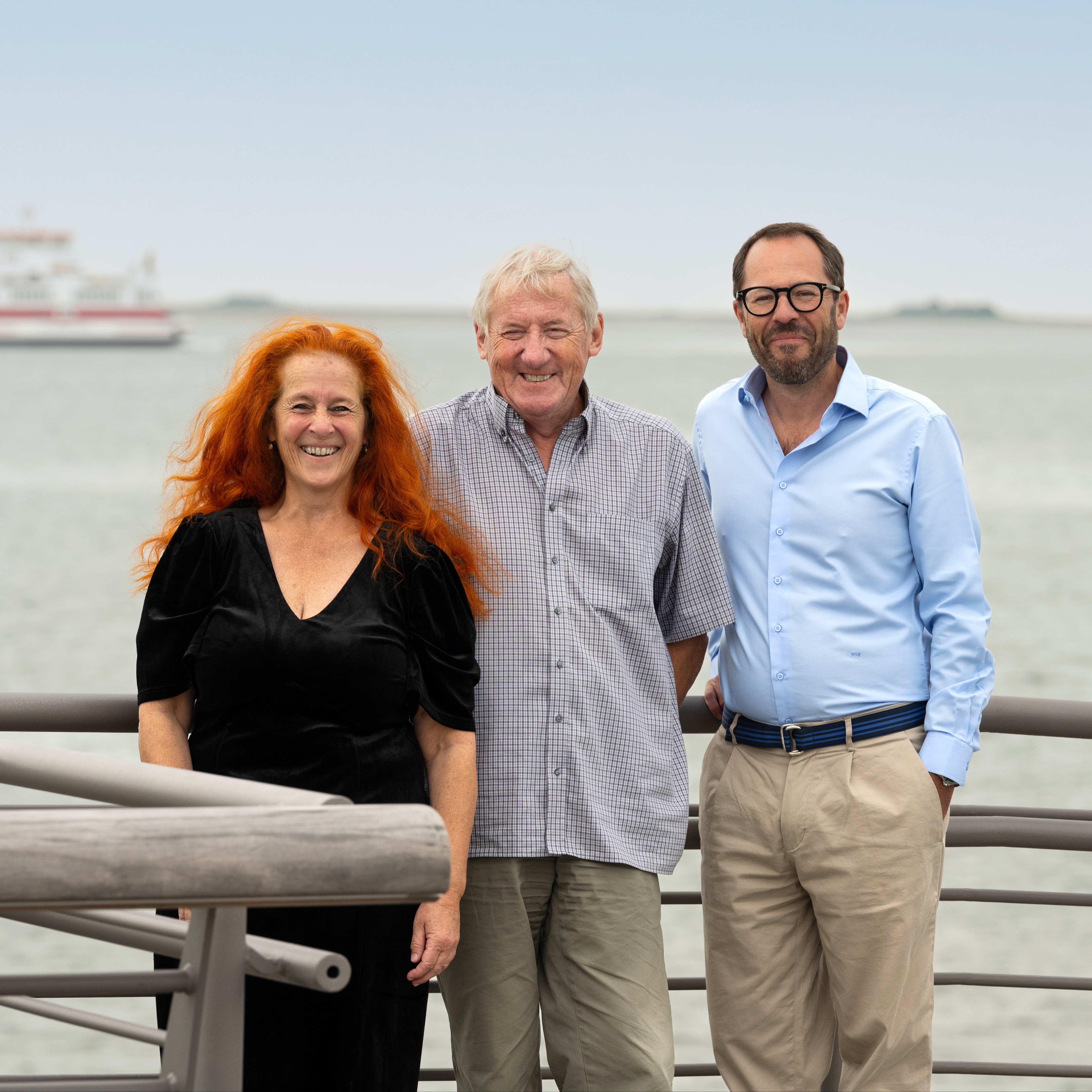 Drei Personen stehen vor einem Geländer mit Wasser und einem Schiff im Hintergrund.