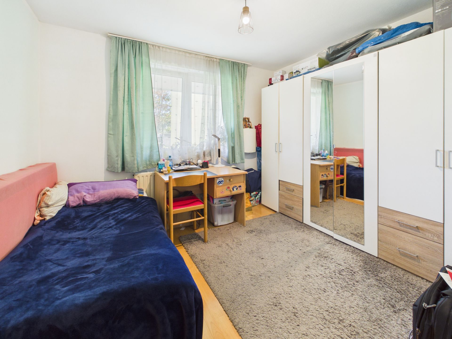 Helles Schlafzimmer mit Einzelbett, Schreibtisch am Fenster und großem Kleiderschrank mit Spiegel.