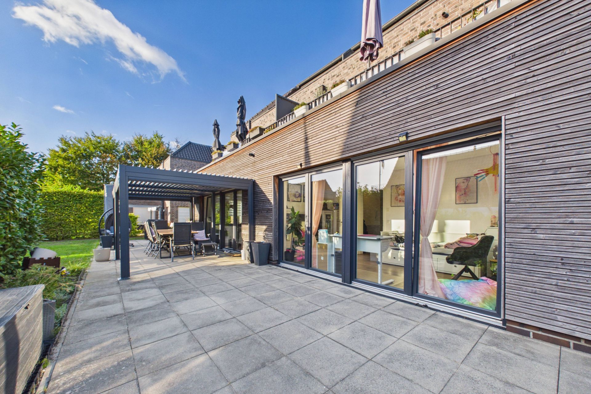 Große Terrasse mit Fliesenboden, angrenzendem Garten und Sitzbereich unter einer Pergola.