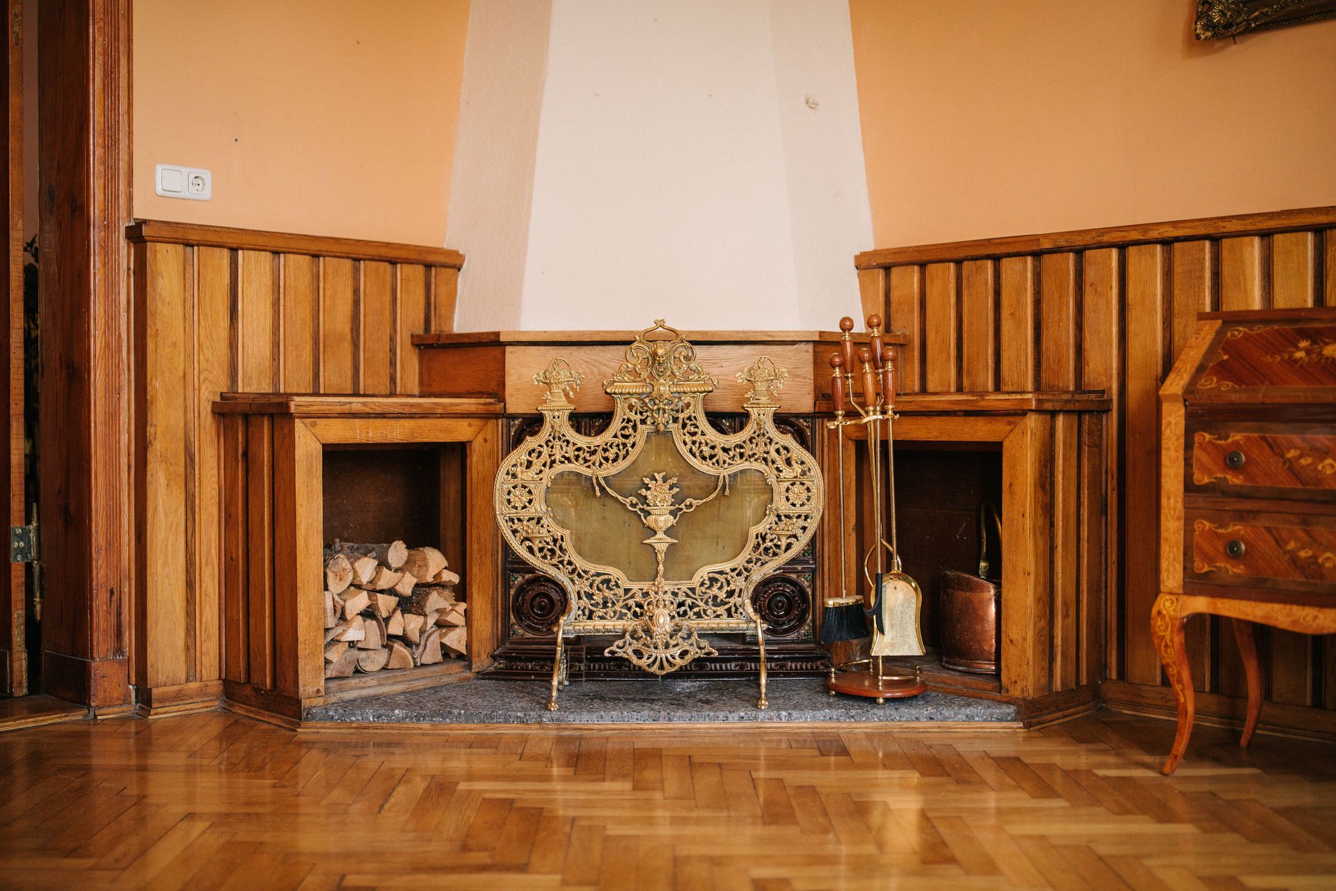 Kamin mit dekorativem Kaminrost, Feuerholz, Kaminbesteck und Holzverkleidung im Wohnraum.