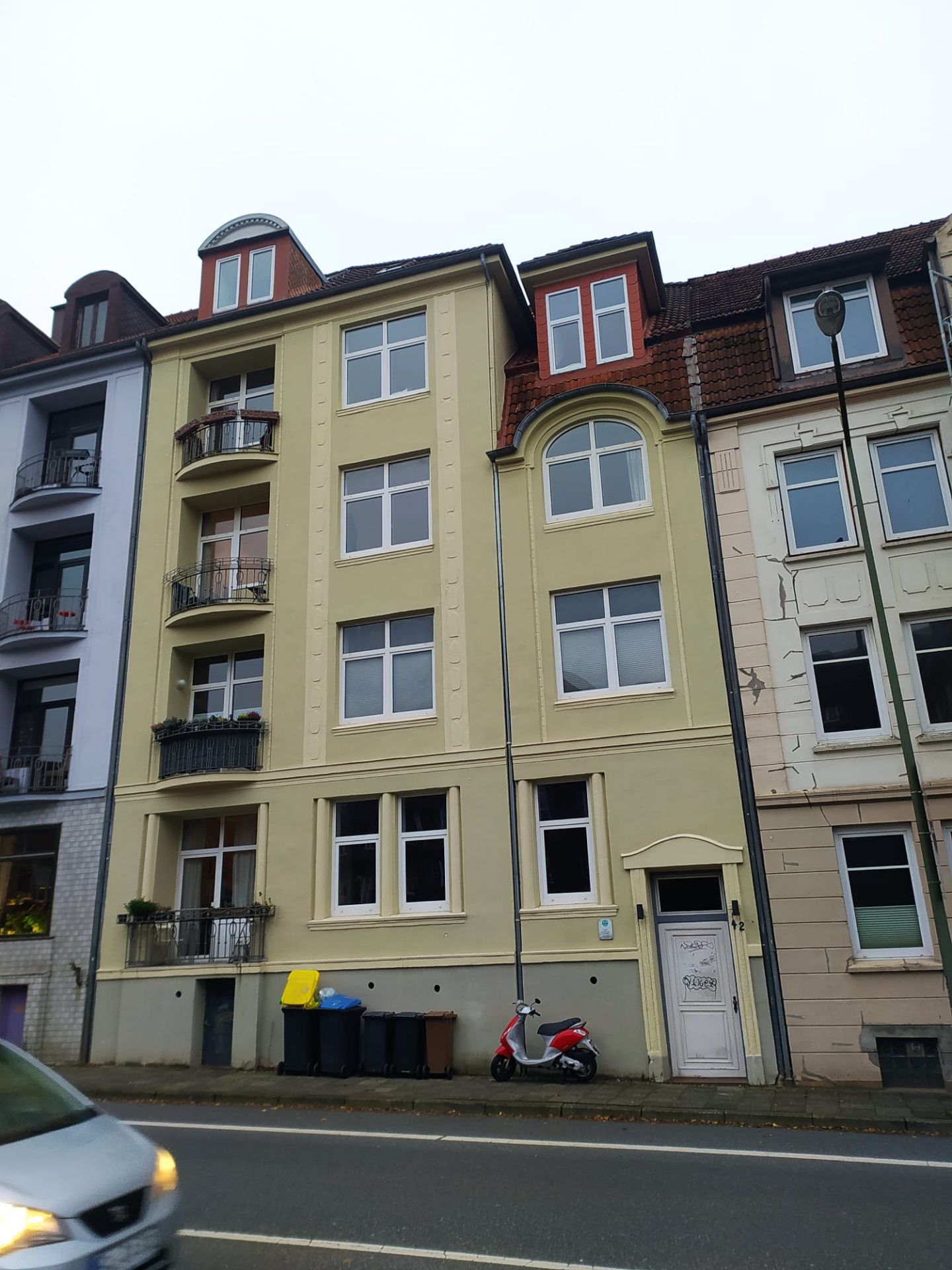 Mehrstöckiges Wohnhaus mit Balkonen und Mülltonnen vor dem Gebäude, ein Roller steht am Gehweg.