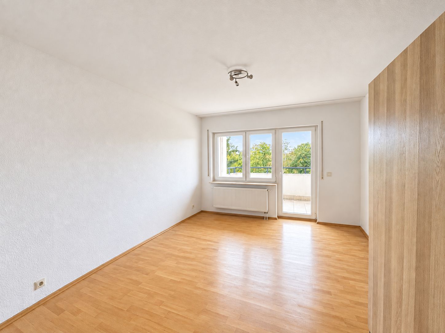 Helles Zimmer mit Holzfußboden, großem Fenster und Tür zum Balkon, weiß gestrichene Wände.