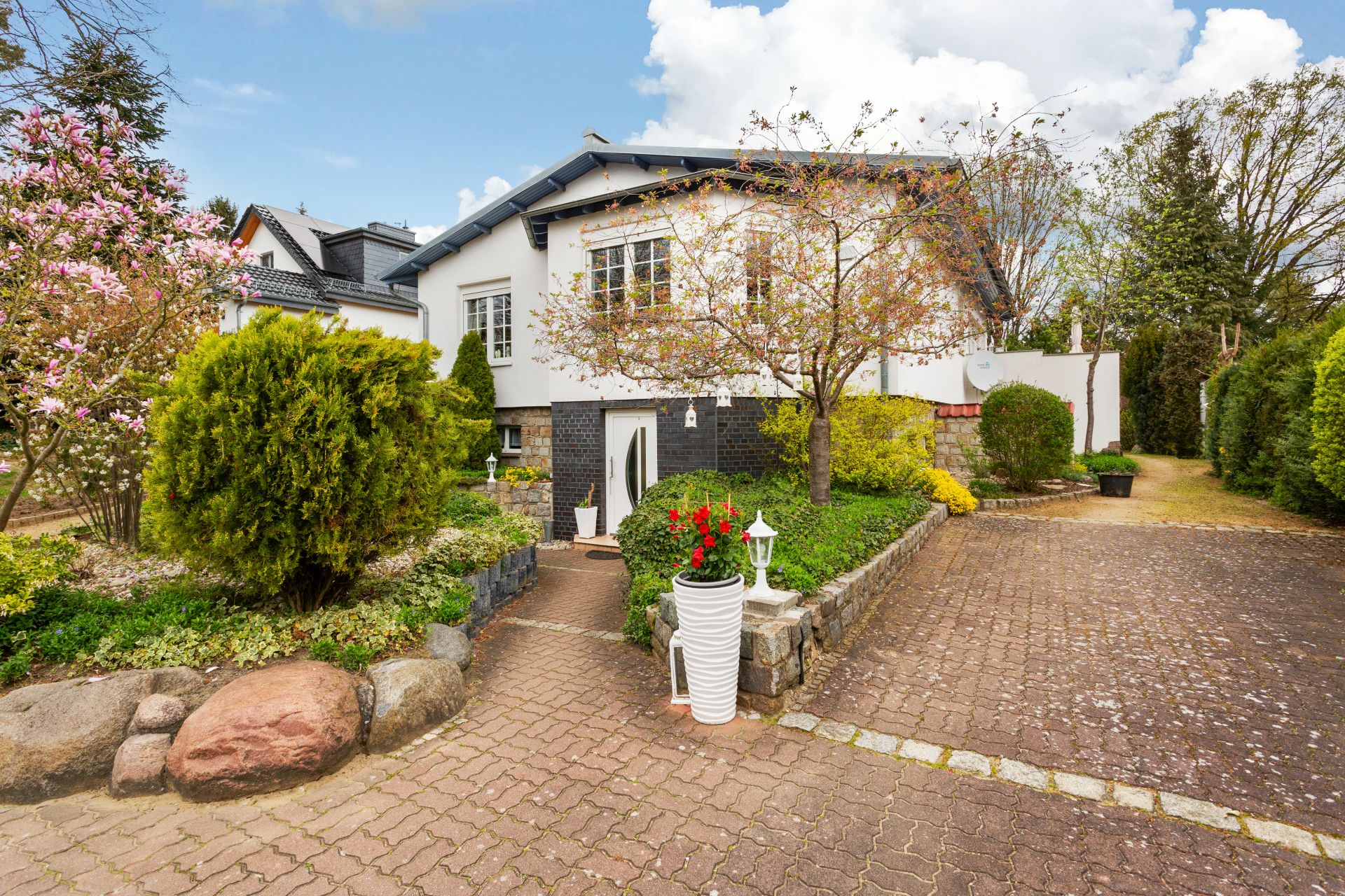 Einfamilienhaus mit Vorgarten, Bäumen, Blumen und gepflasterter Einfahrt.