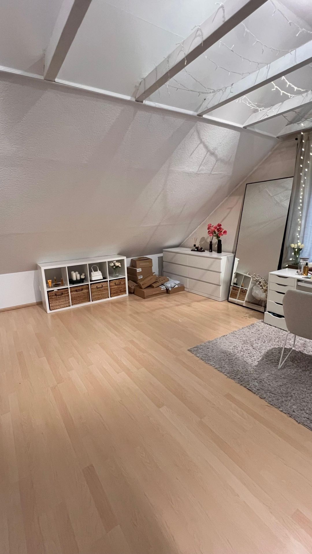 Helles Zimmer mit Dachschräge, Holzfußboden, Kommoden, Spiegel und Regal mit Körben.