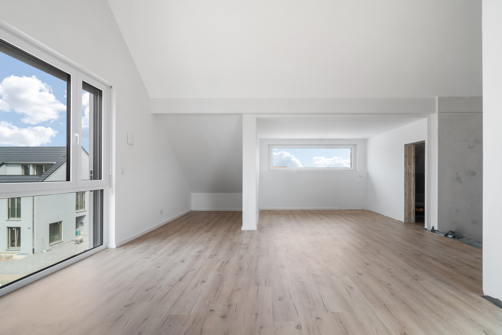 Leerer, heller Raum mit Holzfußboden, großen Fenstern und weiß gestrichenen Wänden.