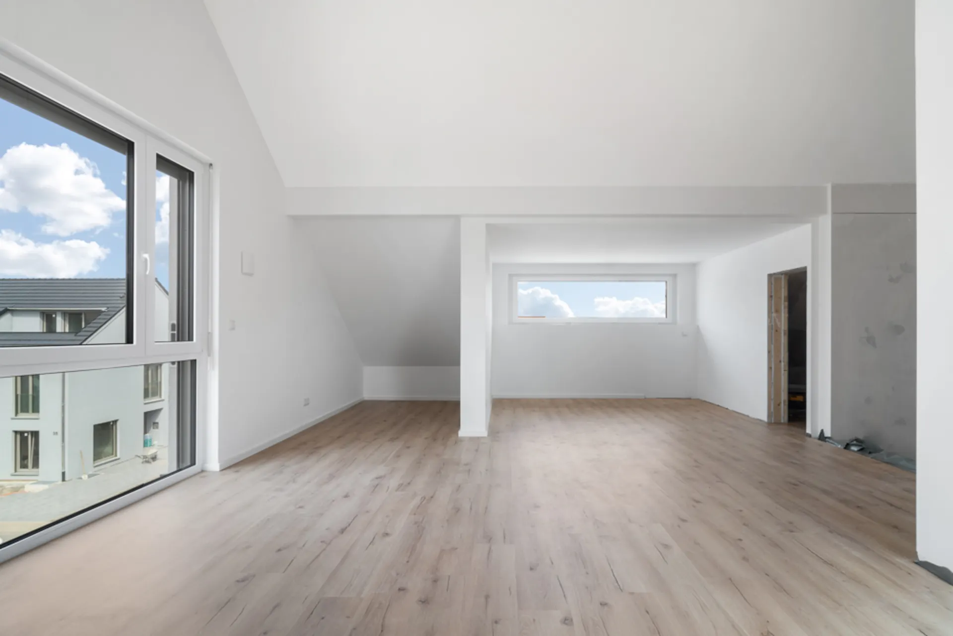 Leerer, heller Raum mit Holzfußboden, großen Fenstern und weiß gestrichenen Wänden.