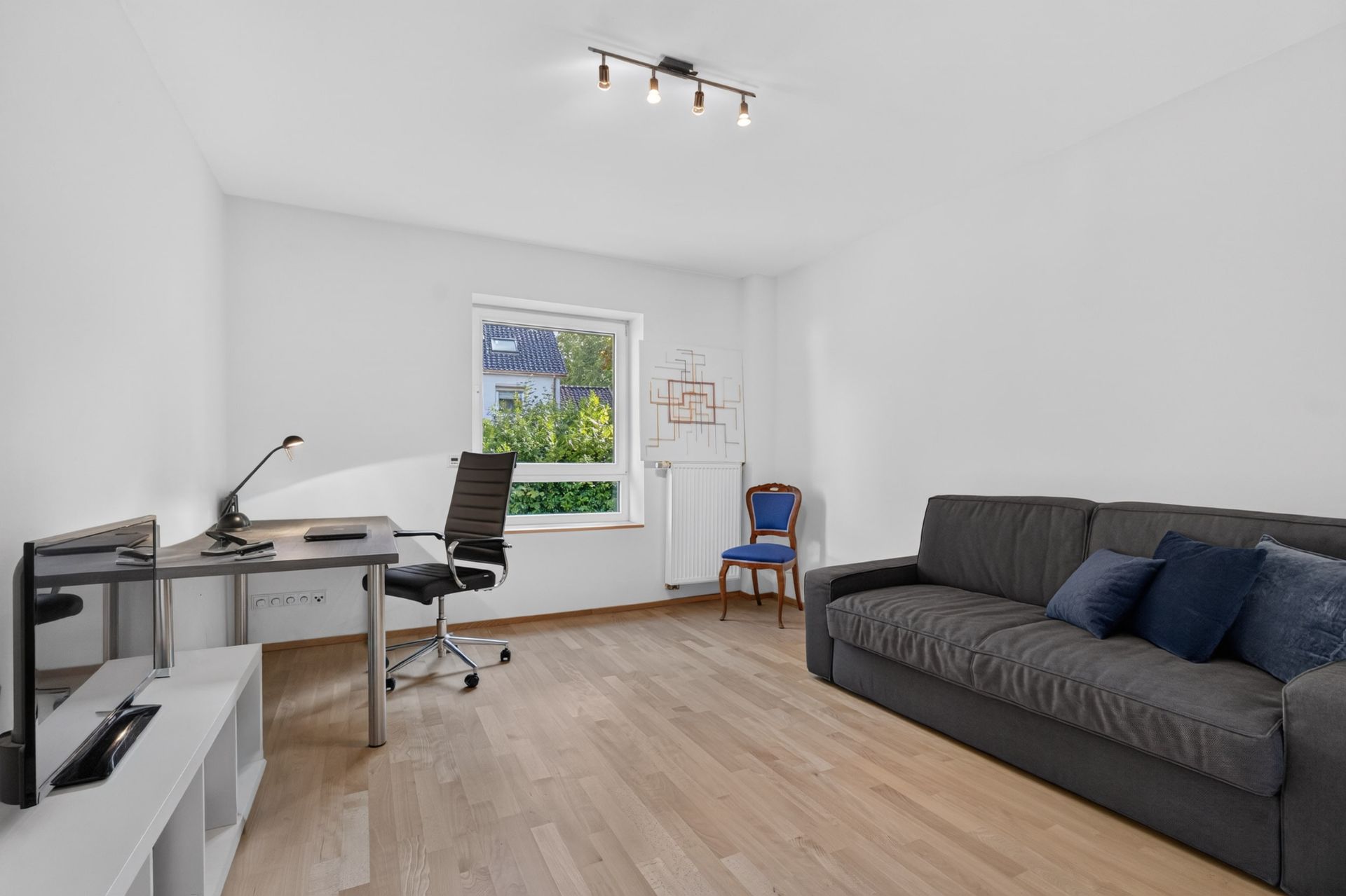 Helles Zimmer mit Schreibtisch, Bürostuhl, Sofa und Fenster, Parkettboden.
