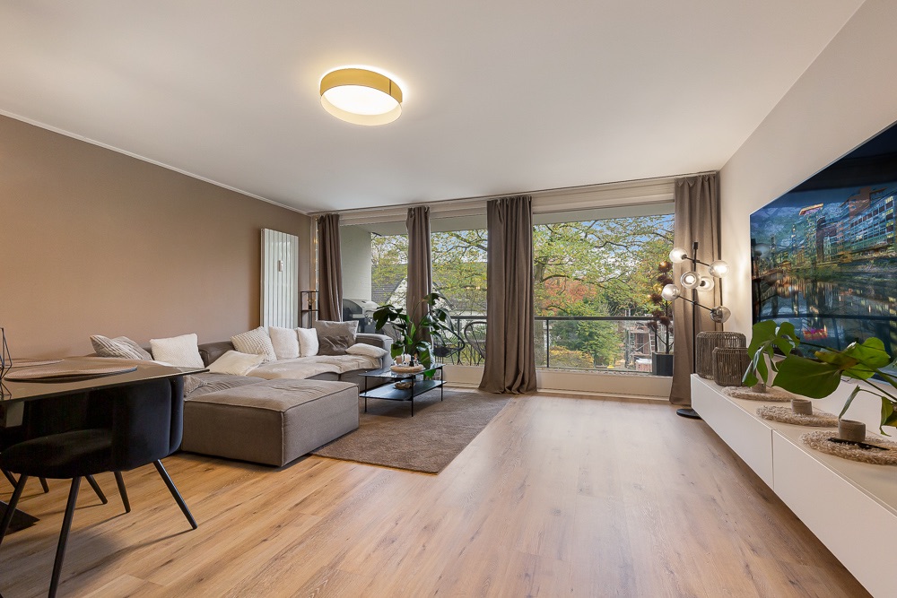 Modernes Wohnzimmer mit Ecksofa, großem Fenster, Holzboden und Pflanzen.