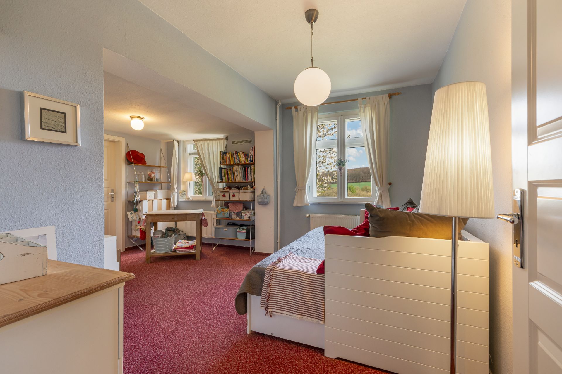 Helles Zimmer mit rotem Teppich, Tagesbett, Stehlampe und Fenster mit Vorhängen.