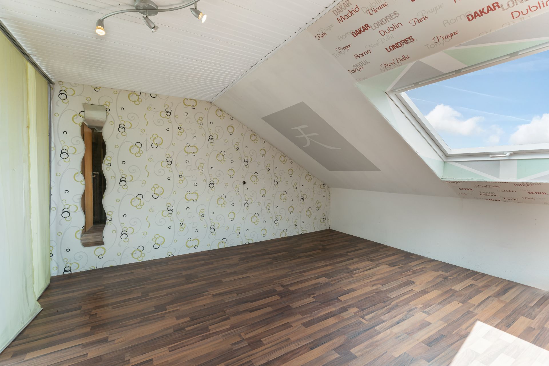 Helles Dachzimmer mit Holzfußboden, gemusterter Tapete und großem Dachfenster.