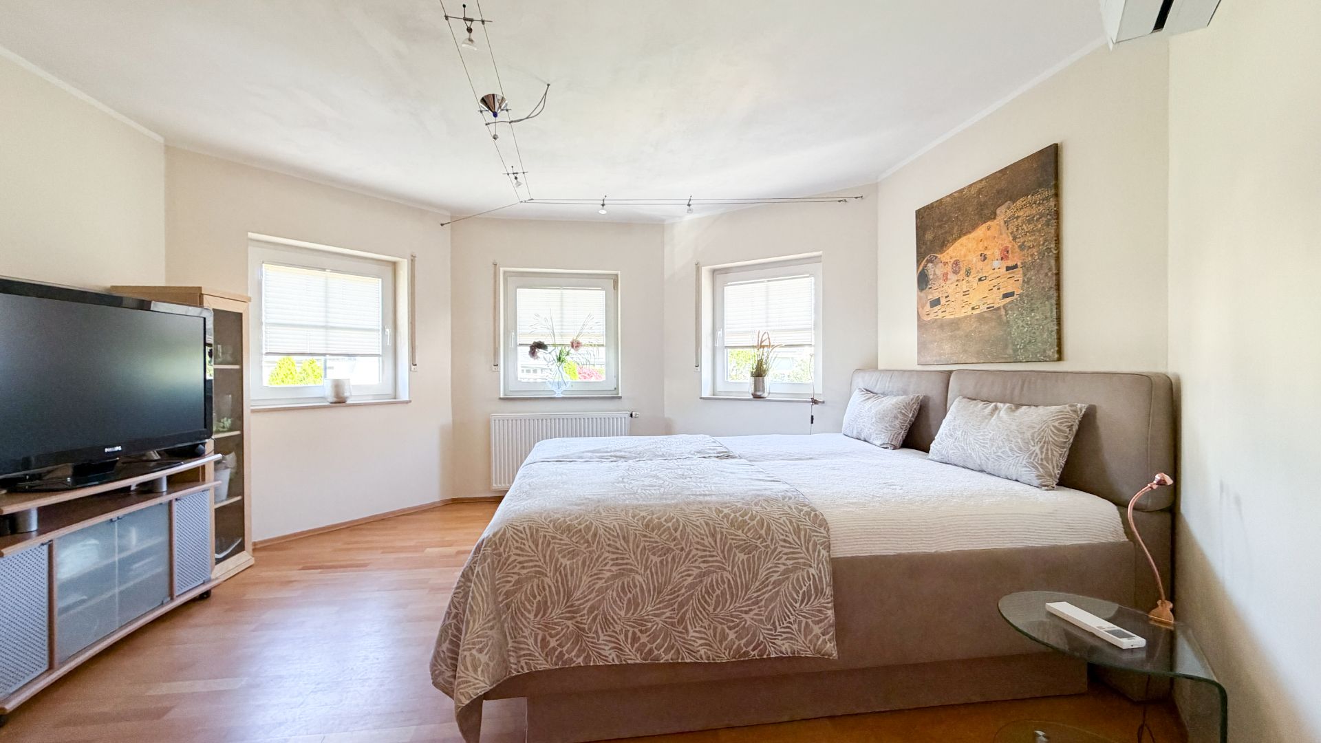Helles Schlafzimmer mit Doppelbett, drei Fenstern, Fernseher und Wandbild.