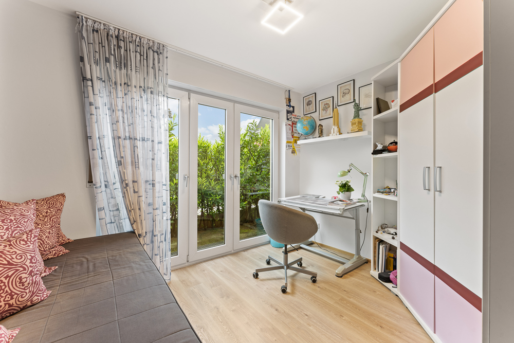 Helles Zimmer mit Schreibtisch, Regal, Kleiderschrank, Schlafcouch und großen Fenstern zum Garten.