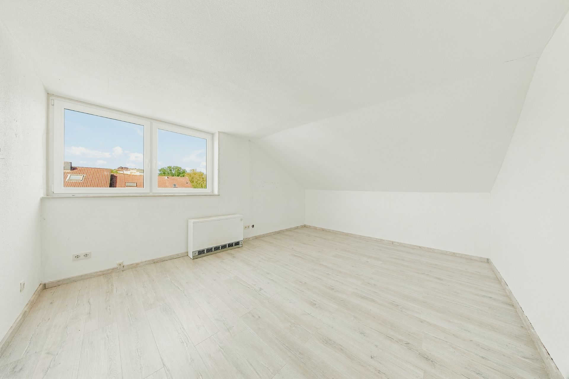 Leerer, heller Raum mit schräger Decke, großem Fenster und hellem Holzboden.