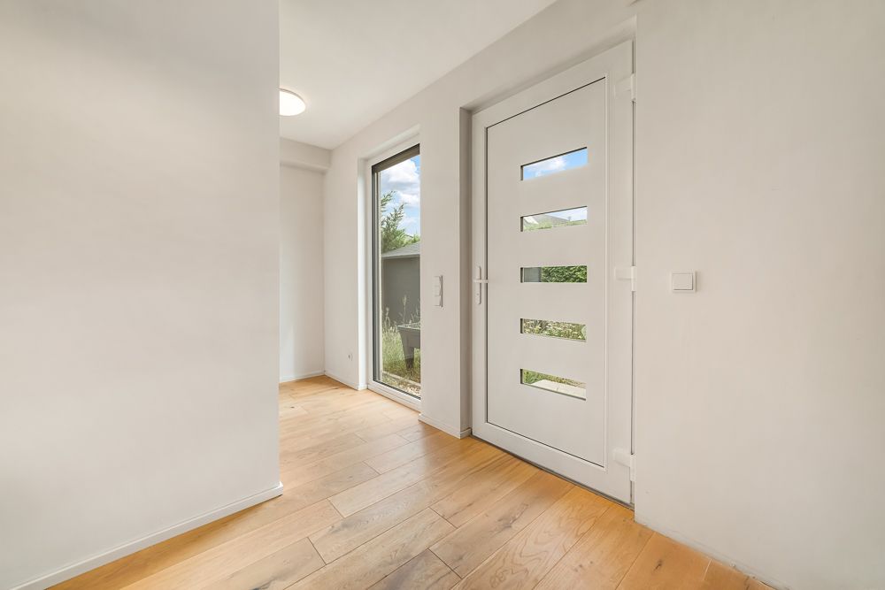 Heller Flur mit weißer Haustür und Holzboden, großes Fenster neben der Tür.