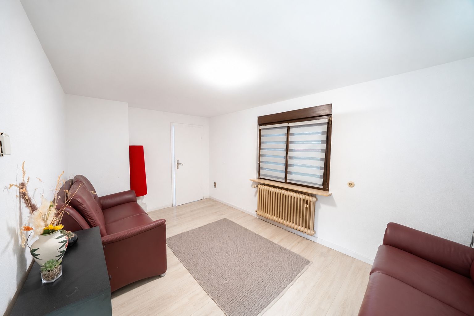 Wohnzimmer mit zwei roten Sofas, Teppich, Fenster mit Jalousien und Heizkörper.