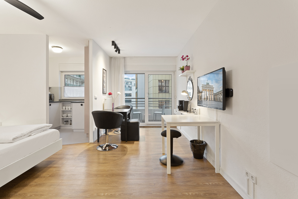 Helles Apartment mit Holzboden, Schreibtisch, Fernseher, Bett und kleinem Balkon.