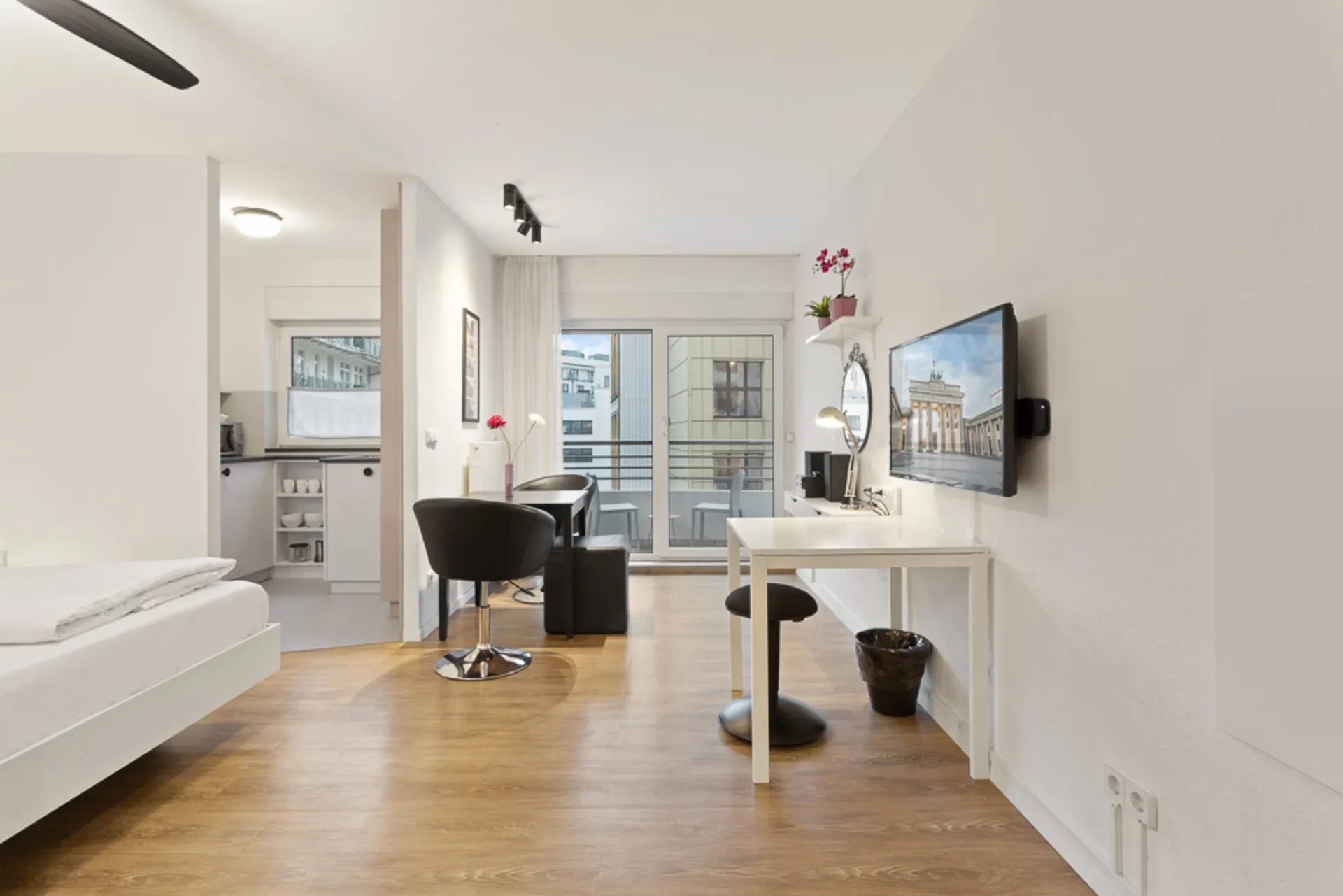 Helles Apartment mit Holzboden, Schreibtisch, Fernseher, Bett und kleinem Balkon.