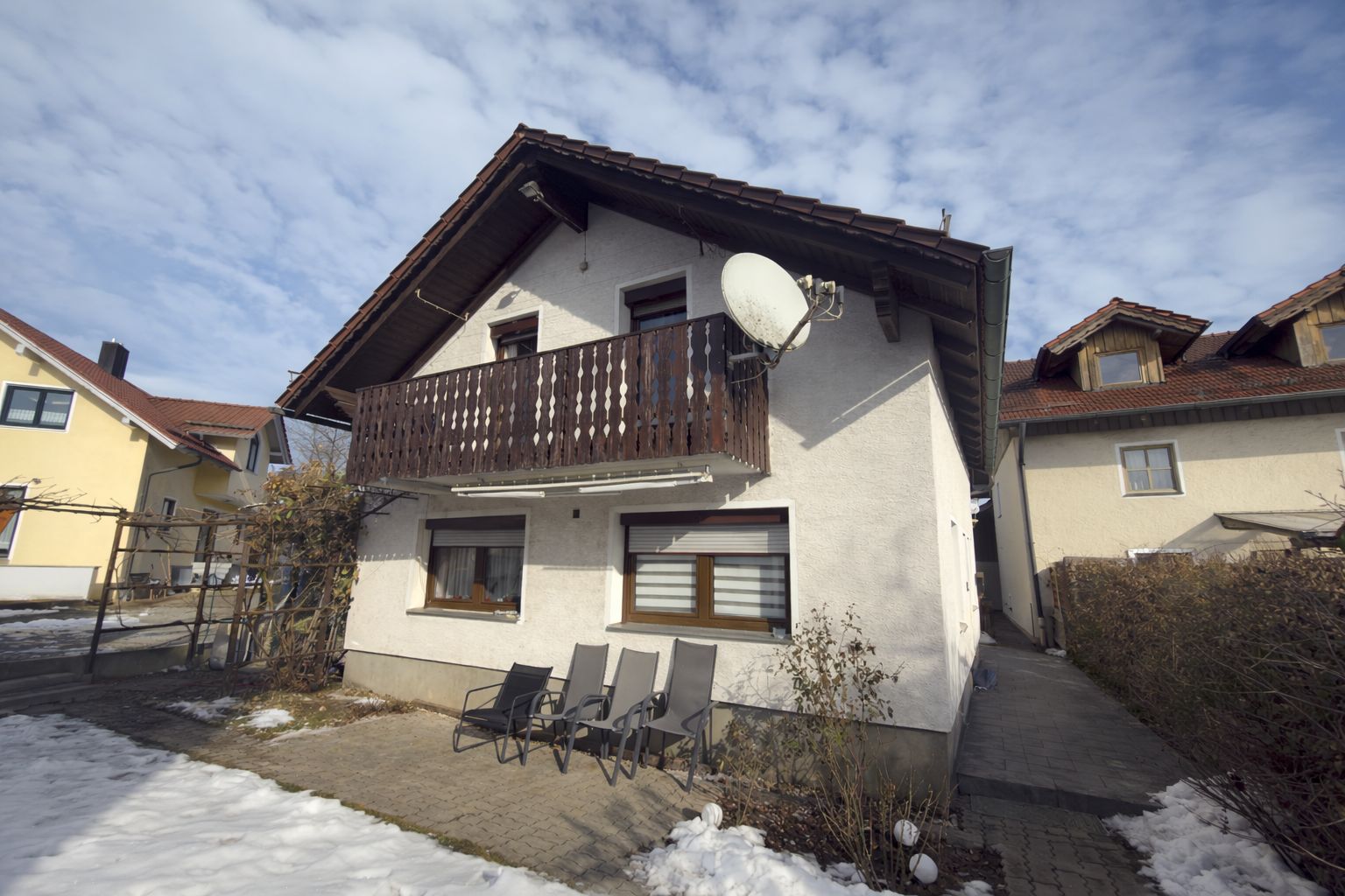 Zweistöckiges Haus mit Balkon, Satellitenschüssel und drei Gartenstühlen vor dem Eingang, teils von Schnee umgeben.