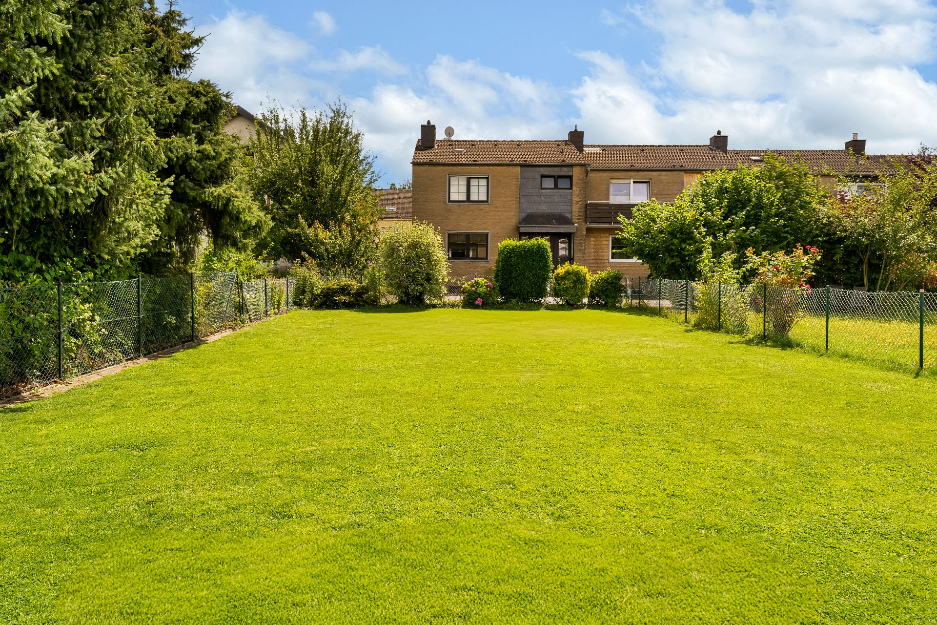Großer, gepflegter Garten mit Rasenfläche, Bäumen und Sträuchern, umgeben von einem Zaun hinter einem Wohnhaus.