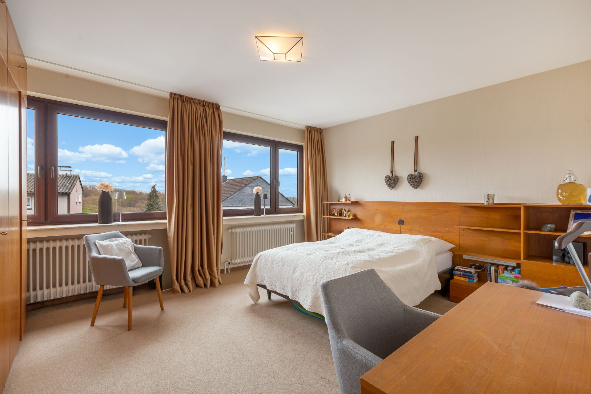 Helles Schlafzimmer mit Doppelbett, zwei Sesseln, Schreibtisch und großen Fenstern mit braunen Vorhängen.