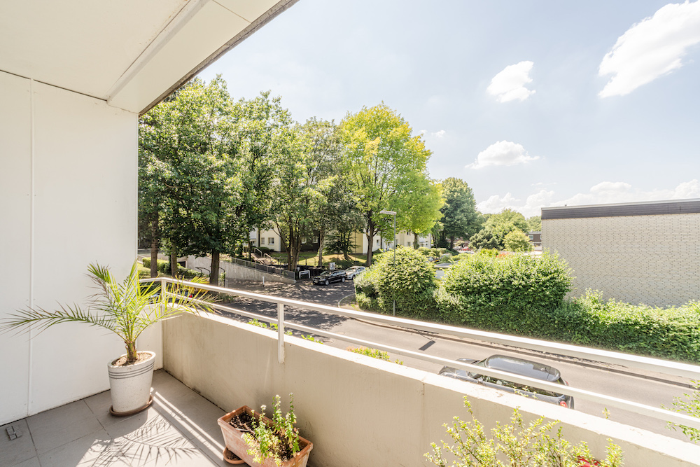 Balkon mit Pflanzen, Blick auf Straße, Bäume und Wohnhäuser bei sonnigem Wetter.