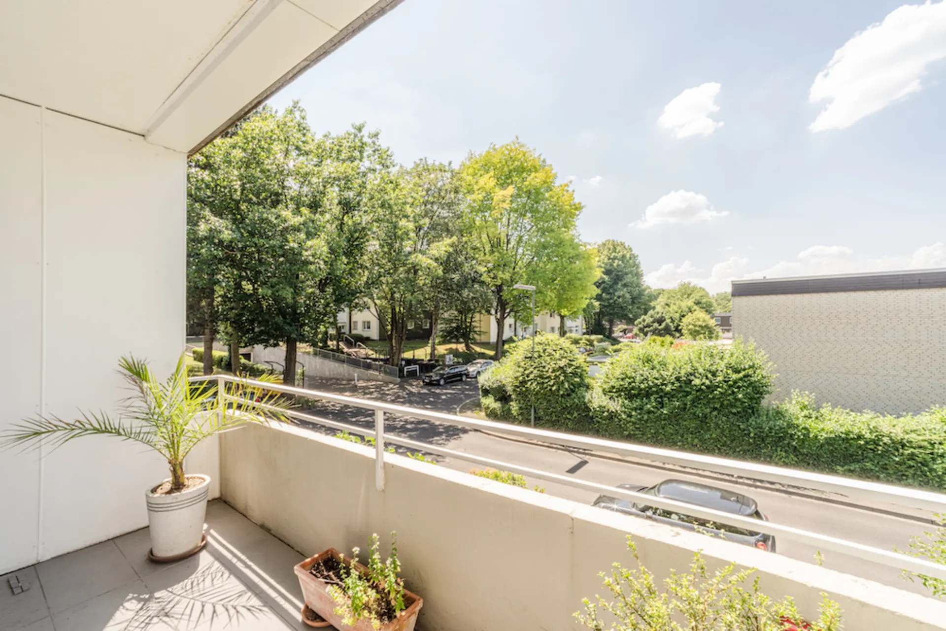 Balkon mit Pflanzen, Blick auf Straße, Bäume und Wohnhäuser bei sonnigem Wetter.