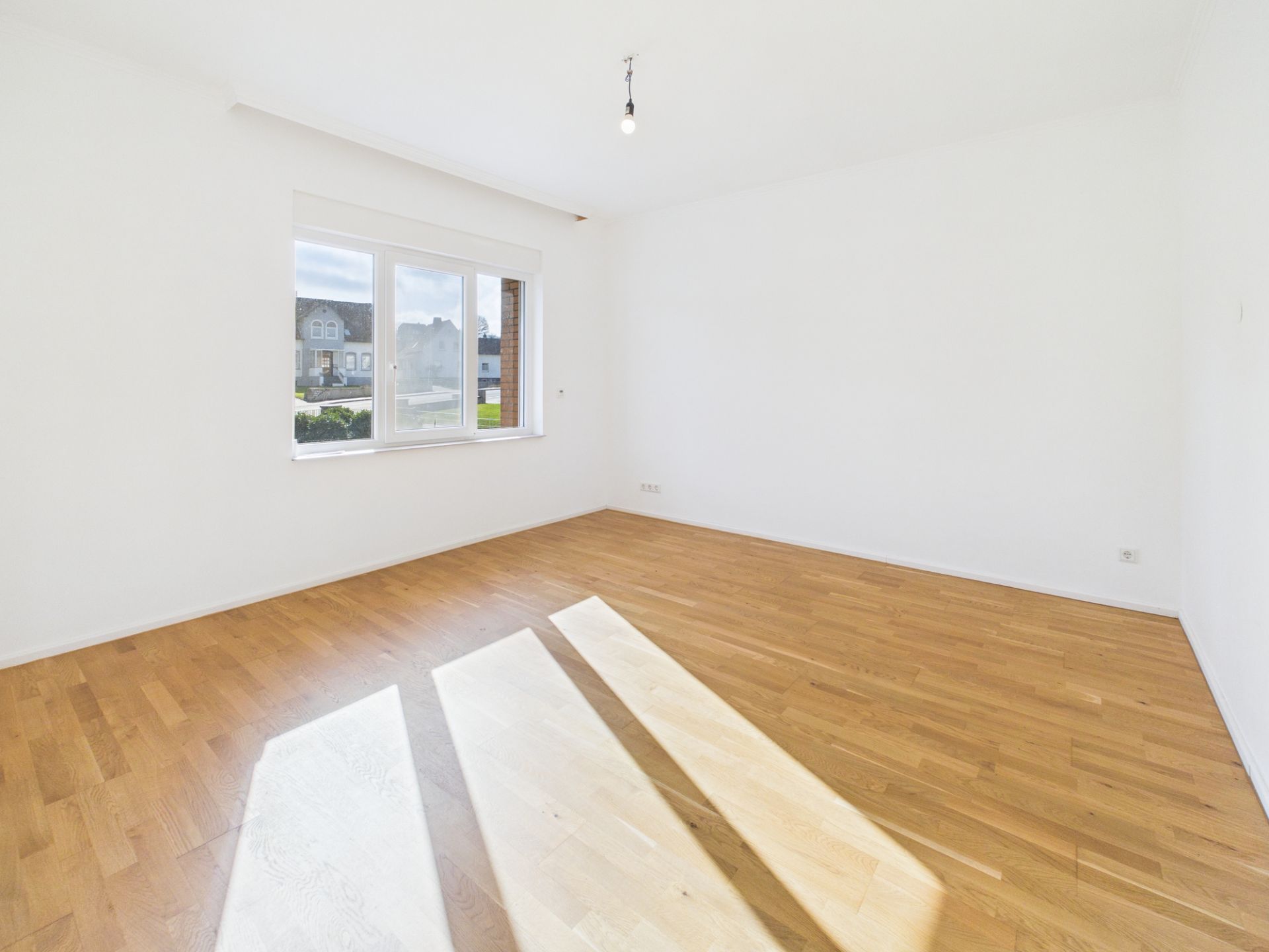 Heller, leerer Raum mit Holzfußboden und großem Fenster, durch das Tageslicht einfällt.